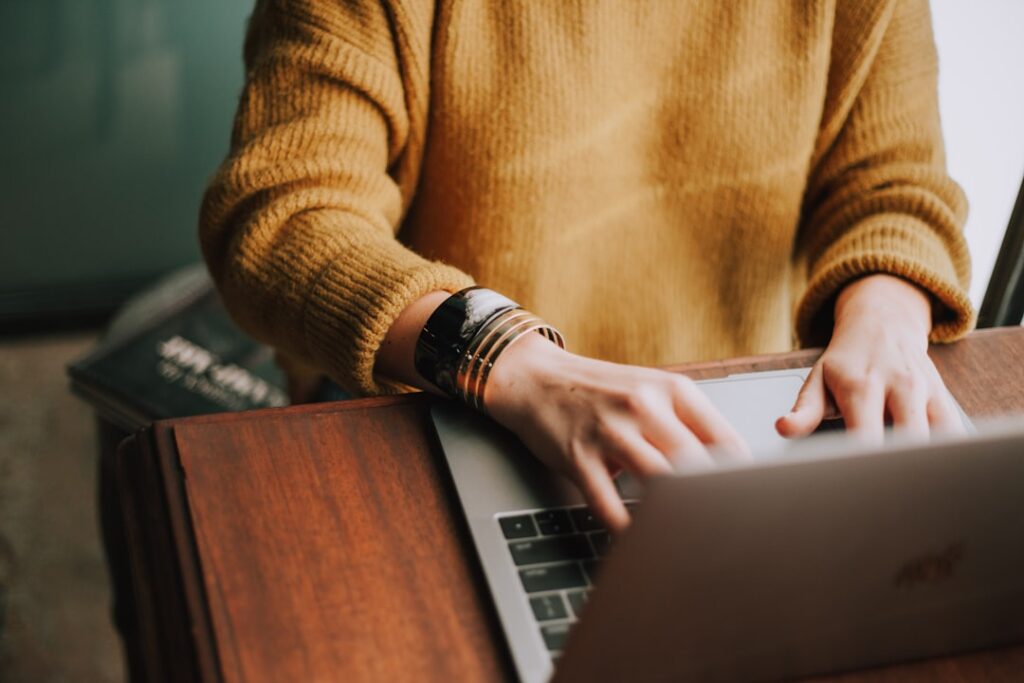 Person in a mustard sweater typing on a laptop at a wooden desk. A stack of books is partially visible in the background. Their focus is on the screen, and they are wearing a bracelet.