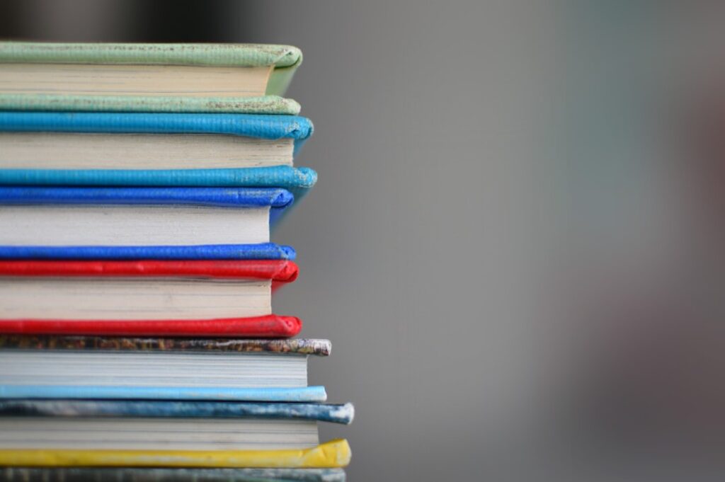 A stack of colorful hardcover books is shown, with covers in various shades of blue, red, yellow, and green. The books are piled unevenly, creating a visually interesting pattern against a blurred gray background.