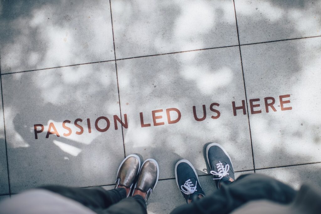 Two pairs of feet stand on a tiled pavement with the words "PASSION LED US HERE" written in bold, capital letters. Shadows of tree leaves are visible on the ground. One pair of shoes is metallic and the other is blue.