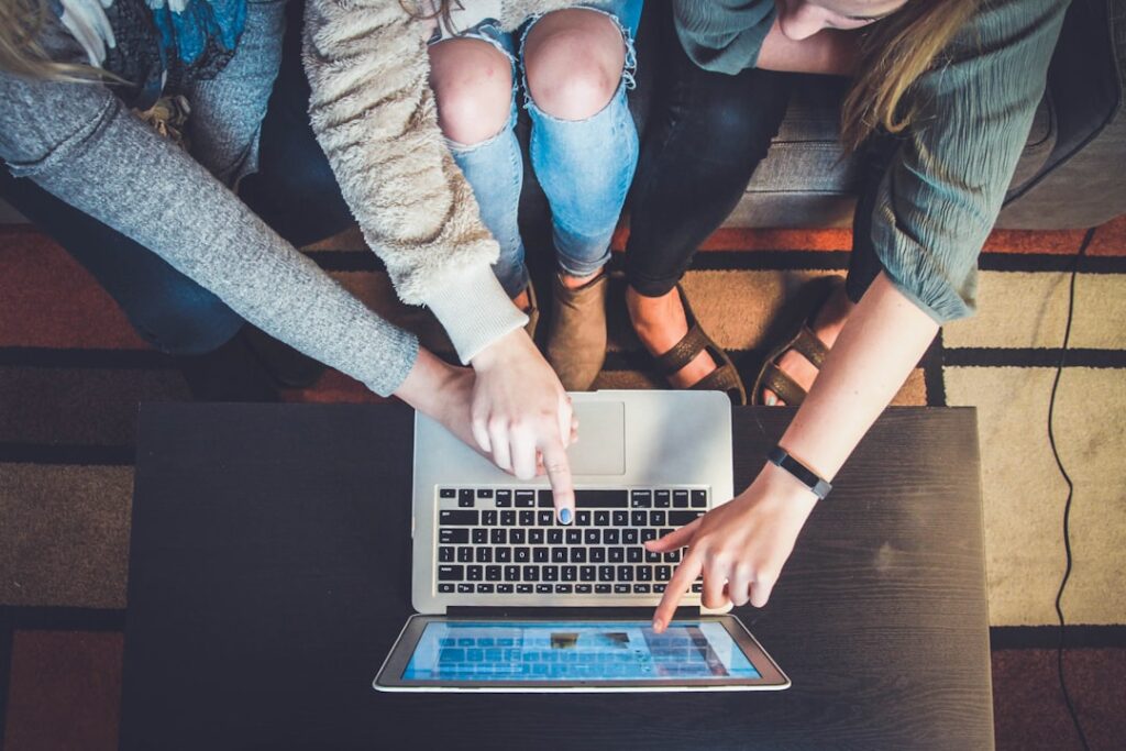 Three people sitting on a couch are collaborating on a laptop. They are pointing at the screen, which displays a blurred image. Casual attire and a wooden coffee table are visible in the setting.