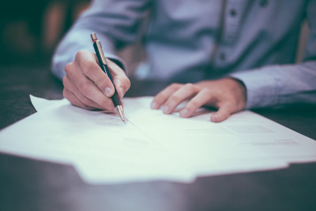 A person in a blue shirt is writing on several papers spread out on a table with a pen in their right hand. The focus is on their hands as they work on the documents. The background is blurred.